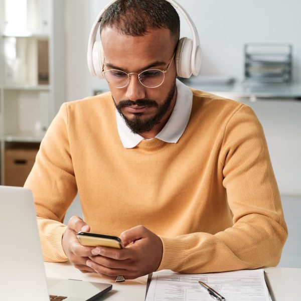 Serious young middle eastern businessman in headphones and eyeglasses sitting at table with papers and checking smartphone