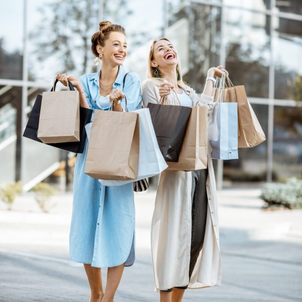 Two happy girlfriends feeling excited with purchases, standing together with shopping bags in front of the shopping mall